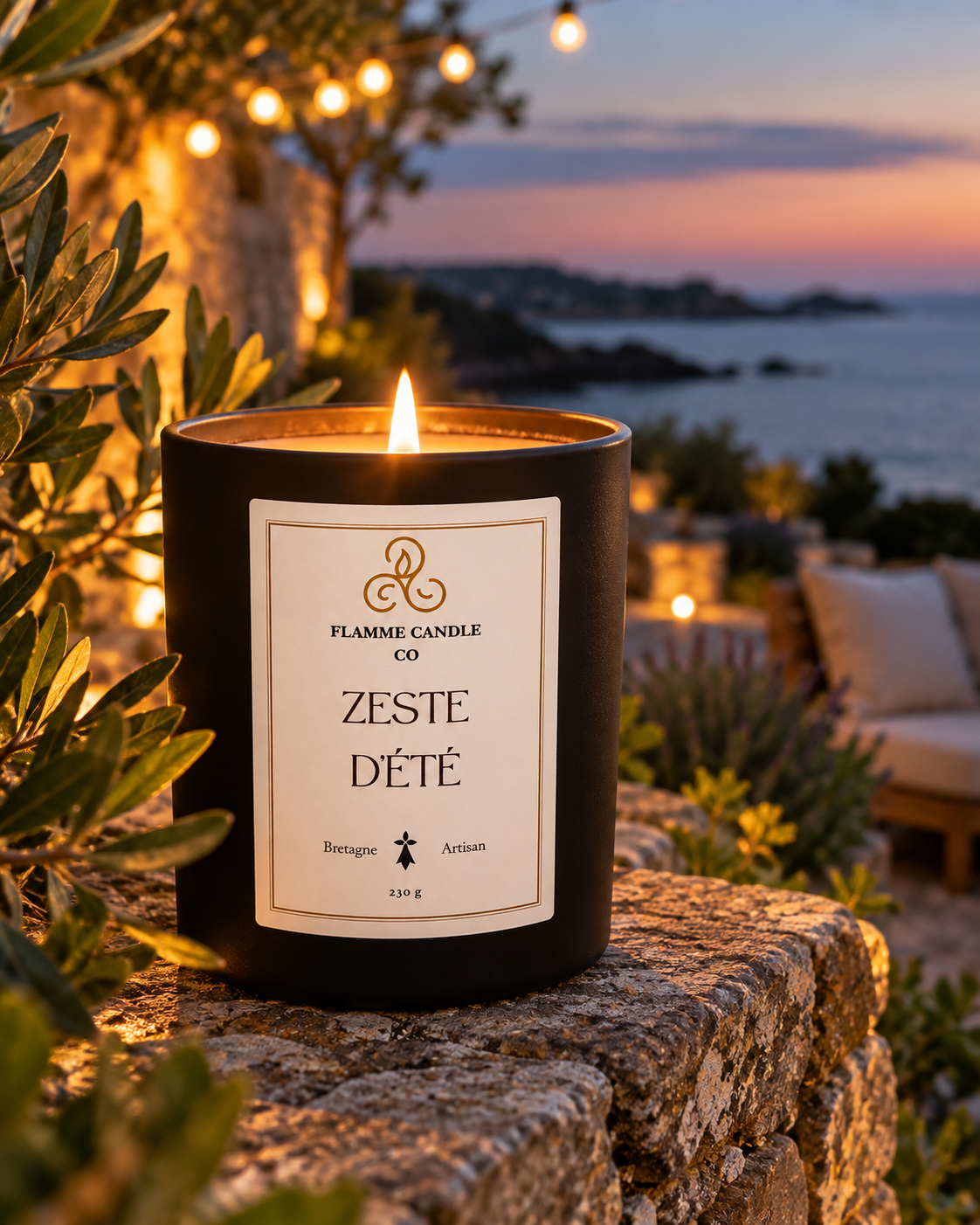Bougie Zeste d'Été allumée sur une pierre entourée de feuillage, avec des guirlandes lumineuses et la mer au crépuscule