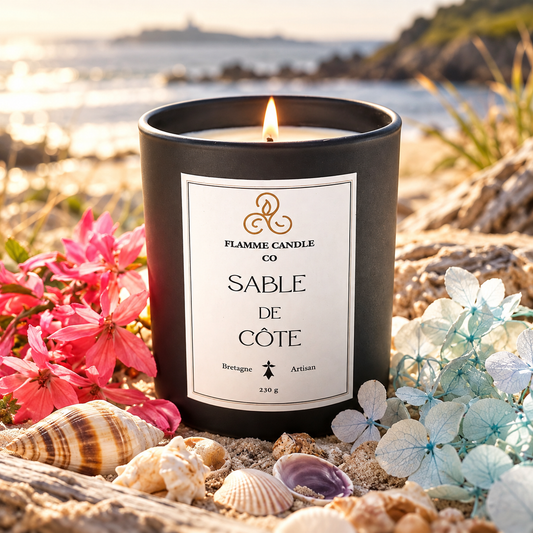 Bougie Sable de Côte allumée sur une plage de sable, entourée de coquillages et de fleurs roses et bleues