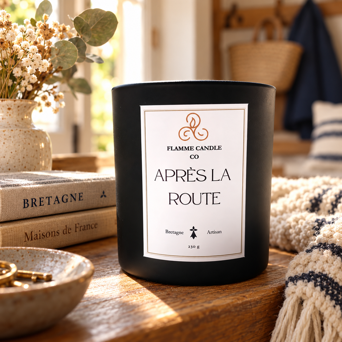 Bougie Après la Route sur une table près d'une pile de livres, d'un bol et d'un plaid rayé