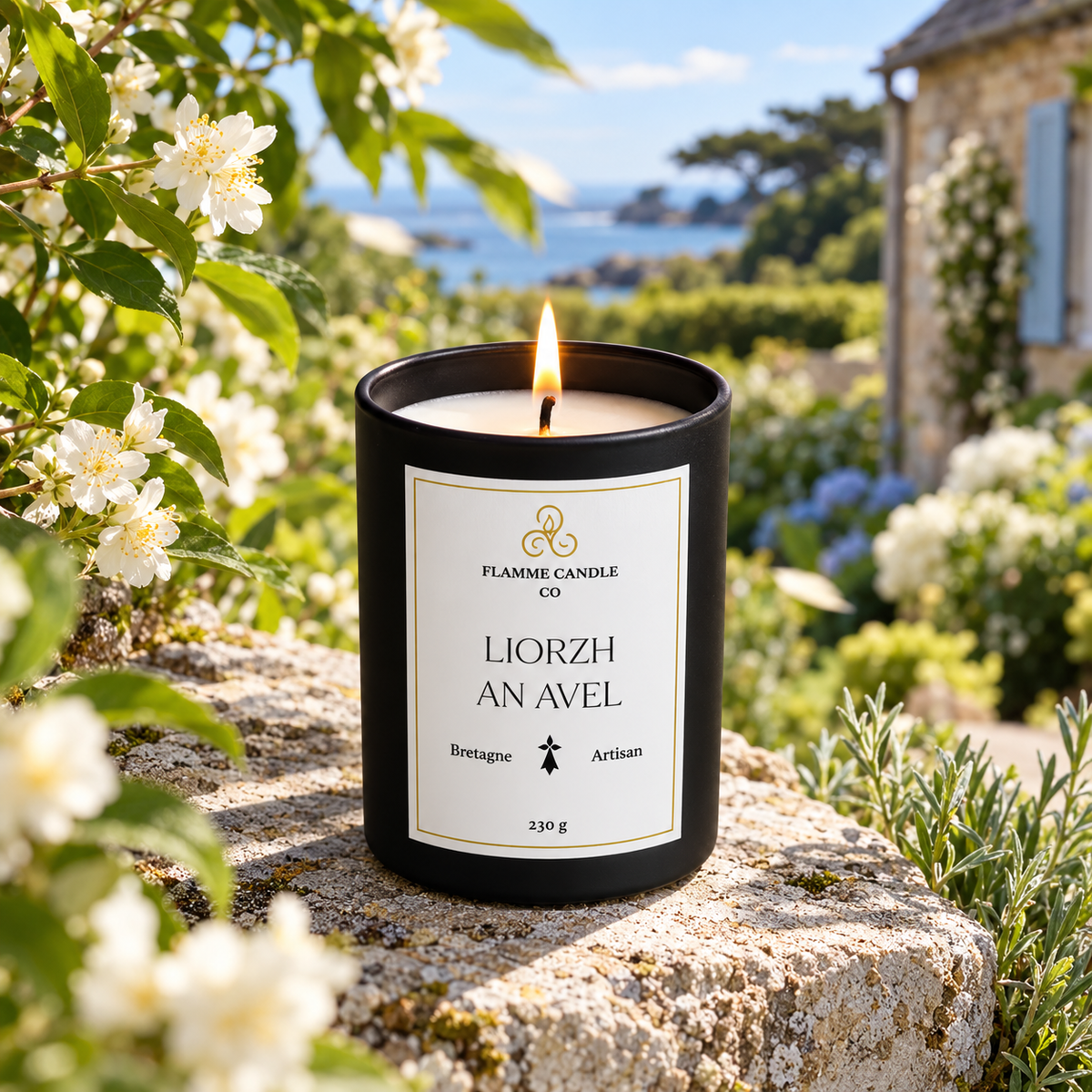 Bougie Liorzh an Avel allumée sur une pierre entourée de fleurs de jasmin blanches, avec une maison bretonne en arrière-plan