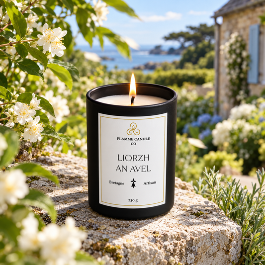 Bougie Liorzh an Avel allumée sur une pierre entourée de fleurs de jasmin blanches, avec une maison bretonne en arrière-plan